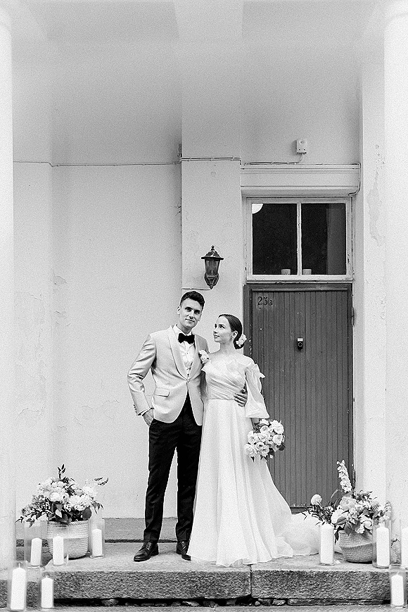 Couple portrait of bride and groom pose on stone steps by a doorway, bride holding bouquet in an off-shoulder gown beside groom in bow tie