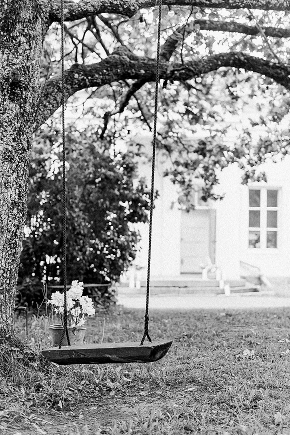 Wedding photo backdrop with a rope swing wooden seat and potted blossoms hanging from a large tree on a lawn by a house porch