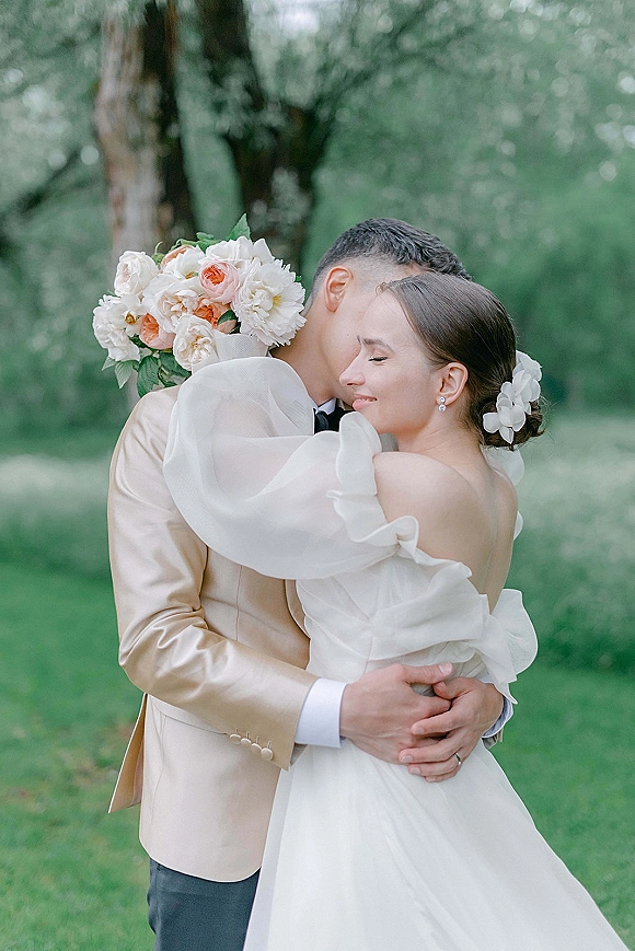 Couple portrait of bride and groom hug, her head resting on his shoulder as she holds a peach rose bouquet on a green lawn with trees