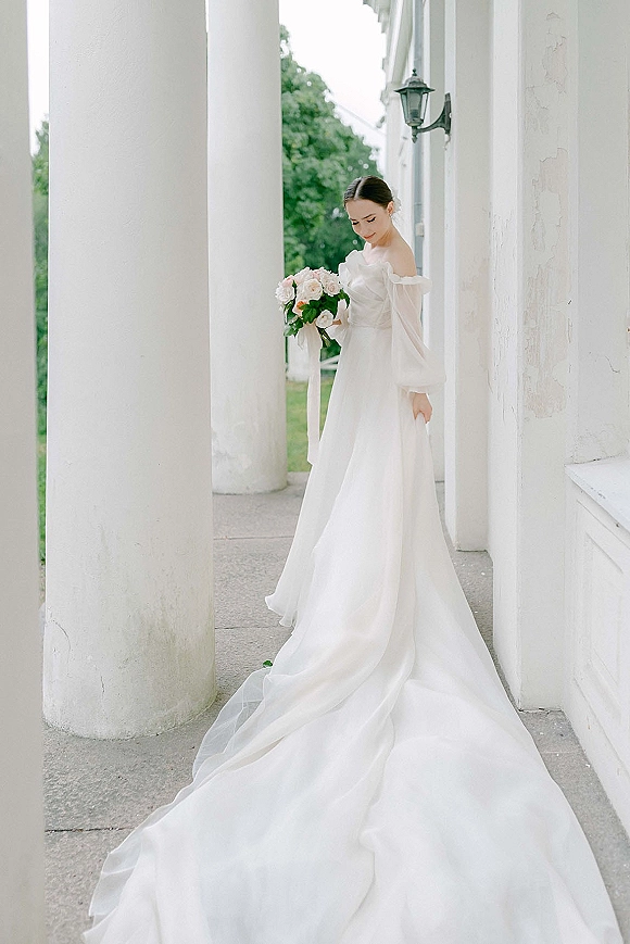 Bridal portrait of a bride looking down, holding a pink rose bouquet with ribbon, her off the shoulder dress and long train on a porch with white columns