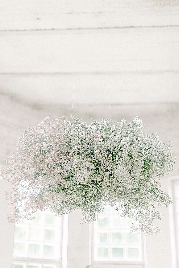 Hanging floral installation of airy baby’s breath suspended by wires, floating like a cloud beneath a white ceiling and window panes