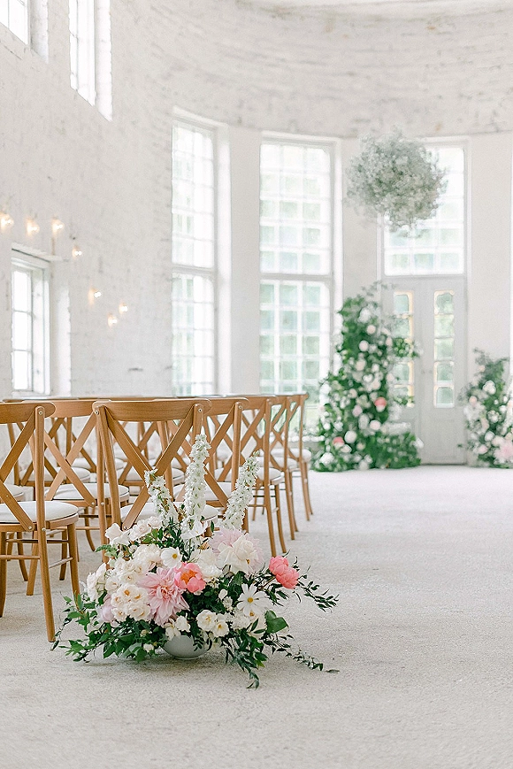 Indoor ceremony setup with an indoor wedding ceremony aisle lined with wood cross-back chairs, blush and white florals, and hanging greenery in a bright white brick room
