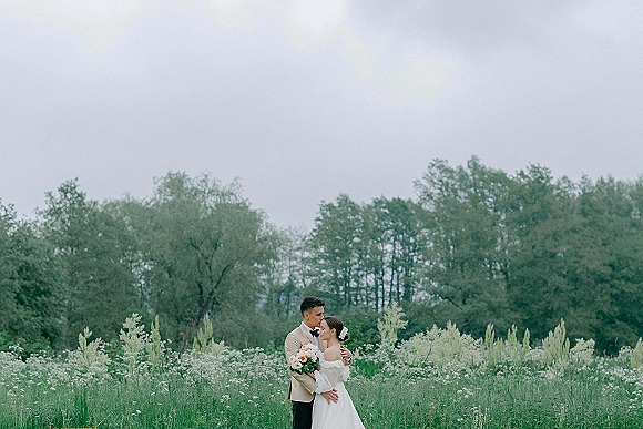Couple portrait of bride and groom embracing, holding a white rose bouquet in a wildflower meadow under an overcast sky
