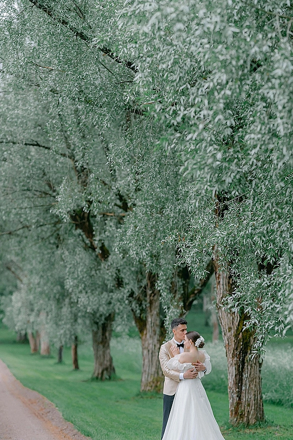 Couple portrait of bride in off-the-shoulder wedding dress and groom in beige tuxedo sharing a forehead kiss on a tree-lined path