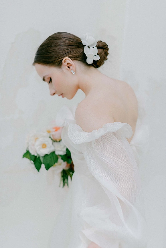 Bridal portrait of a bride looking down in an off shoulder wedding dress with orchid hair pins, holding a white and peach bouquet against a white wall