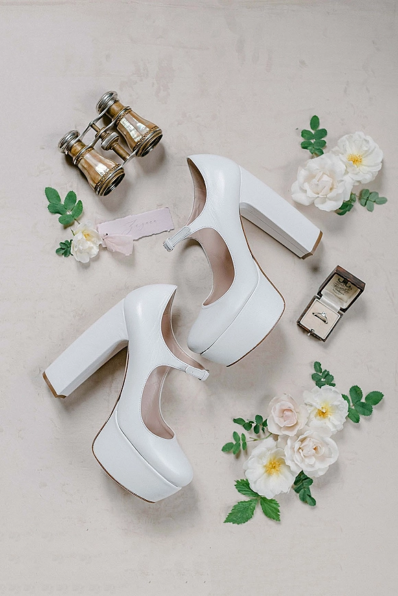 Wedding shoes flatlay with white platform wedding heels, ring box and band, vintage binoculars, petals and greenery on neutral fabric backdrop