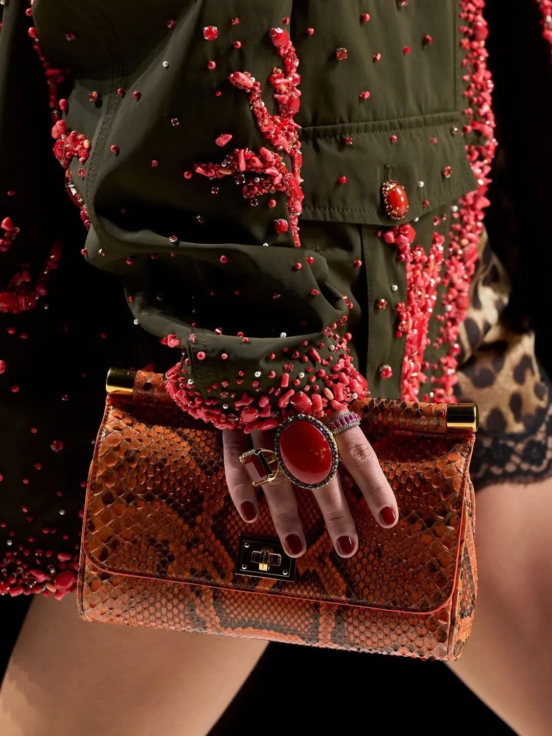 Hand with a red cocktail ring holding a snakeskin top-handle bag against a beaded olive jacket