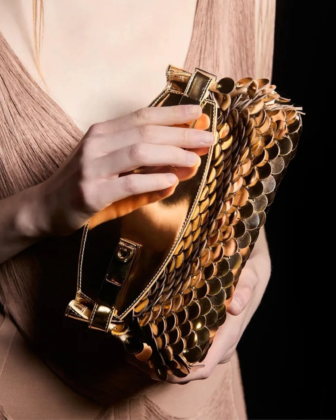Sculptural gold scale-effect clutch held against a blush pleated dress
