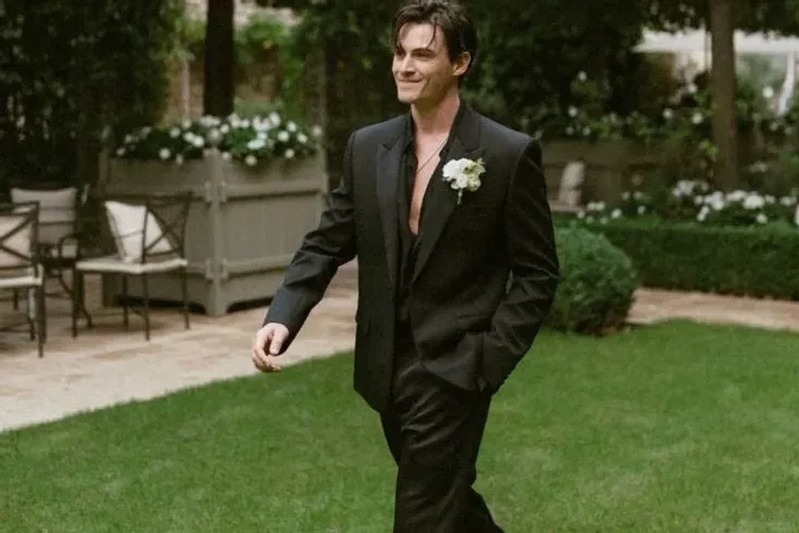 A groom walking wearing a dark suit with an unbuttoned black collared shirt underneath and a floral boutonniere.