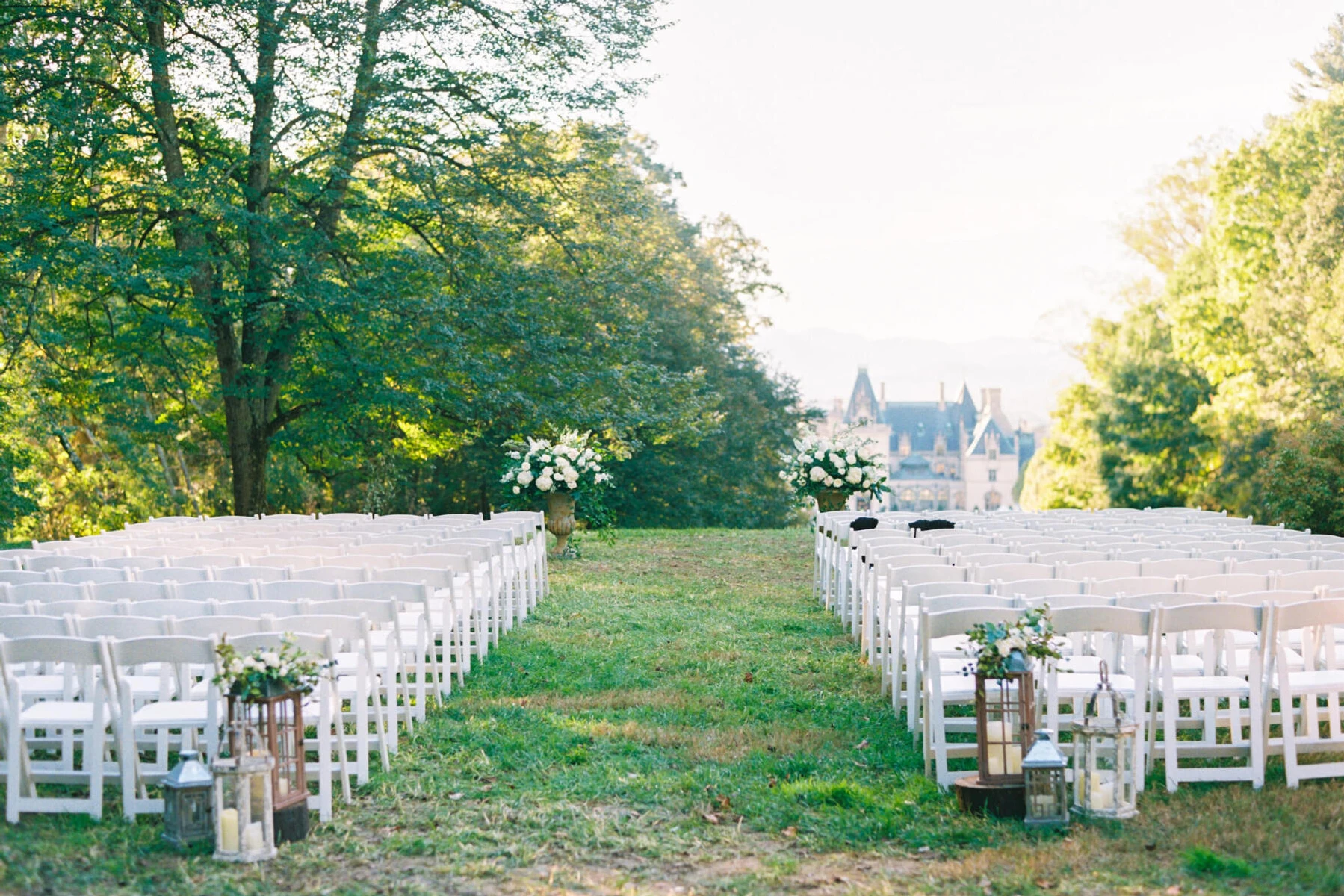 Castle Wedding Venues: An outdoor ceremony setup on a lawn with trees around it and a distant castle-like property in the background.