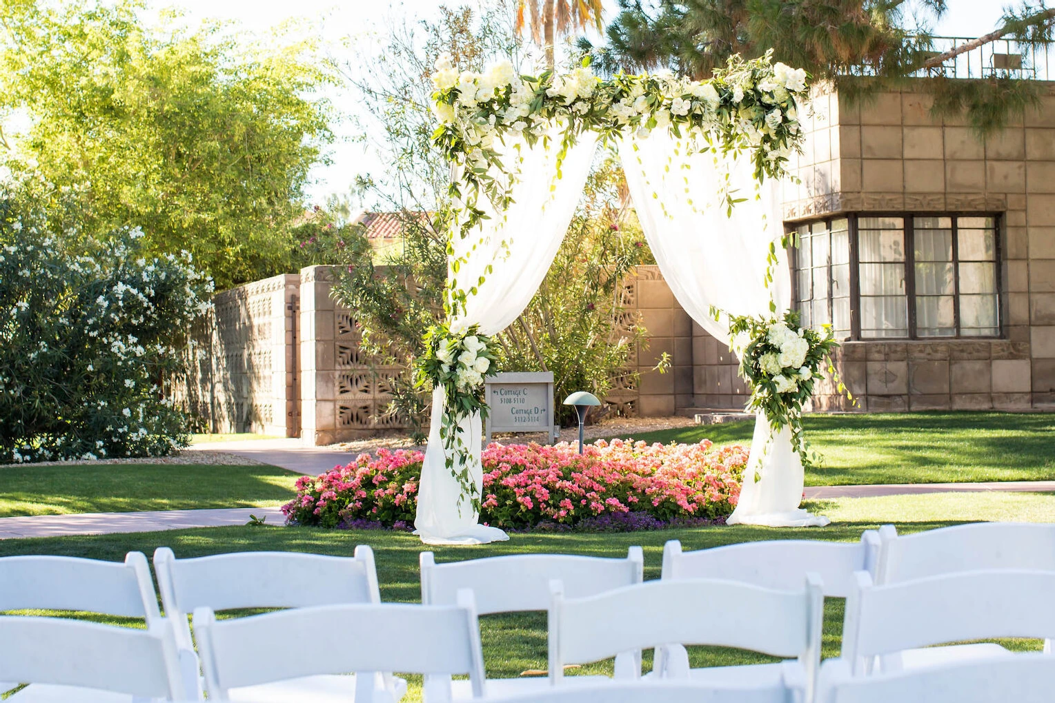 Celebrity Wedding: An outdoor wedding ceremony in a garden area in Arizona, complete with white chairs and an arch with drapery and greenery covering it.