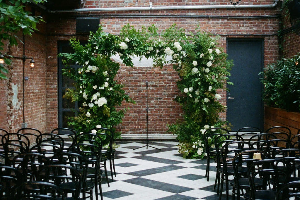 Celebrity Wedding: An indoor ceremony with an arch of greenery and white flowers, black chairs, and black-and-white checkered flooring.