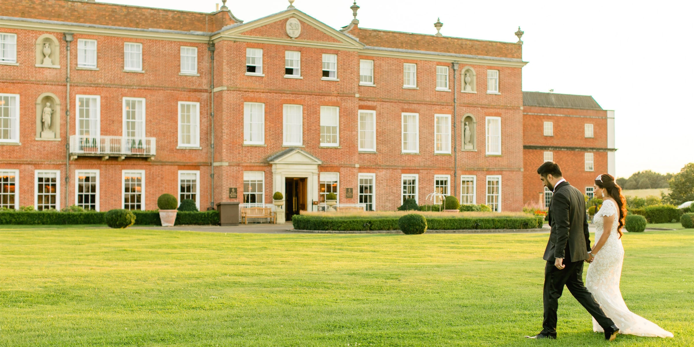 A groom and bride stroll hand-in-hand on the grounds of the Four Seasons Hampshire after their colorful countryside wedding ceremony in England.