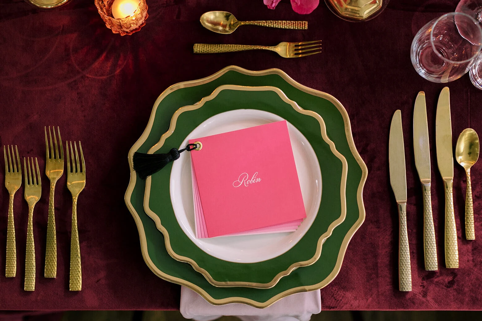 A placesetting, with multiple pieces of gold flatware for the multi-meal dinner reception, flank a stack of fluted green plates and a pink menu with a tassel on it.