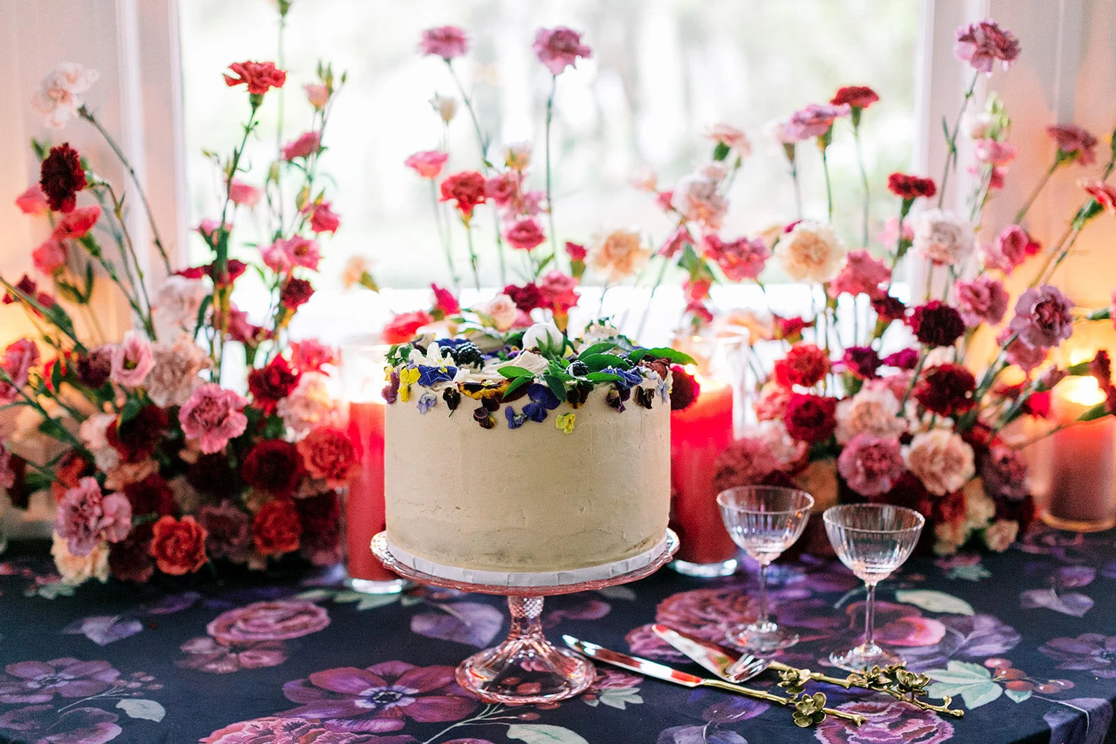 A one-tier wedding cake covered with edible flowers sits on a floral table cloth and is surrounded by fresh flowers at a colorful wedding in North Carolina.