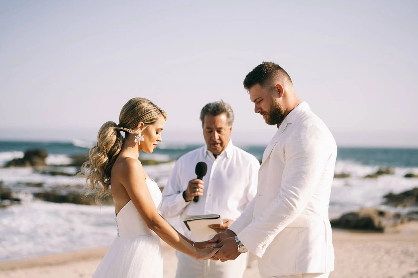 A bride, groom, and their officiant, all wearing white, partake in a glam beach wedding in Mexico.