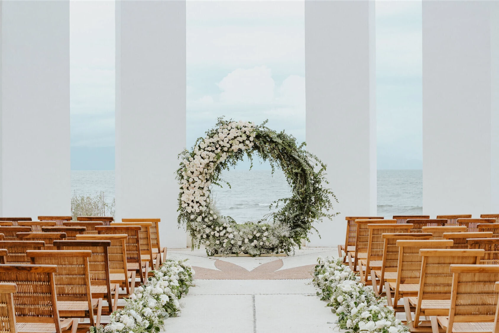 Grand Velas Riviera Nayarit: A circle-shaped floral arch at the end of an aisle, with wooden chairs and white floral arrangements on either side.