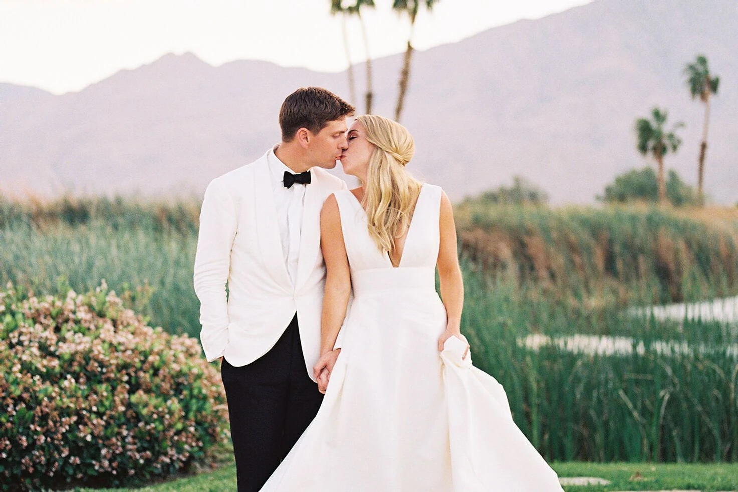 Groom in white jacket tuxedo groom attire posing with bride in a waterfront garden