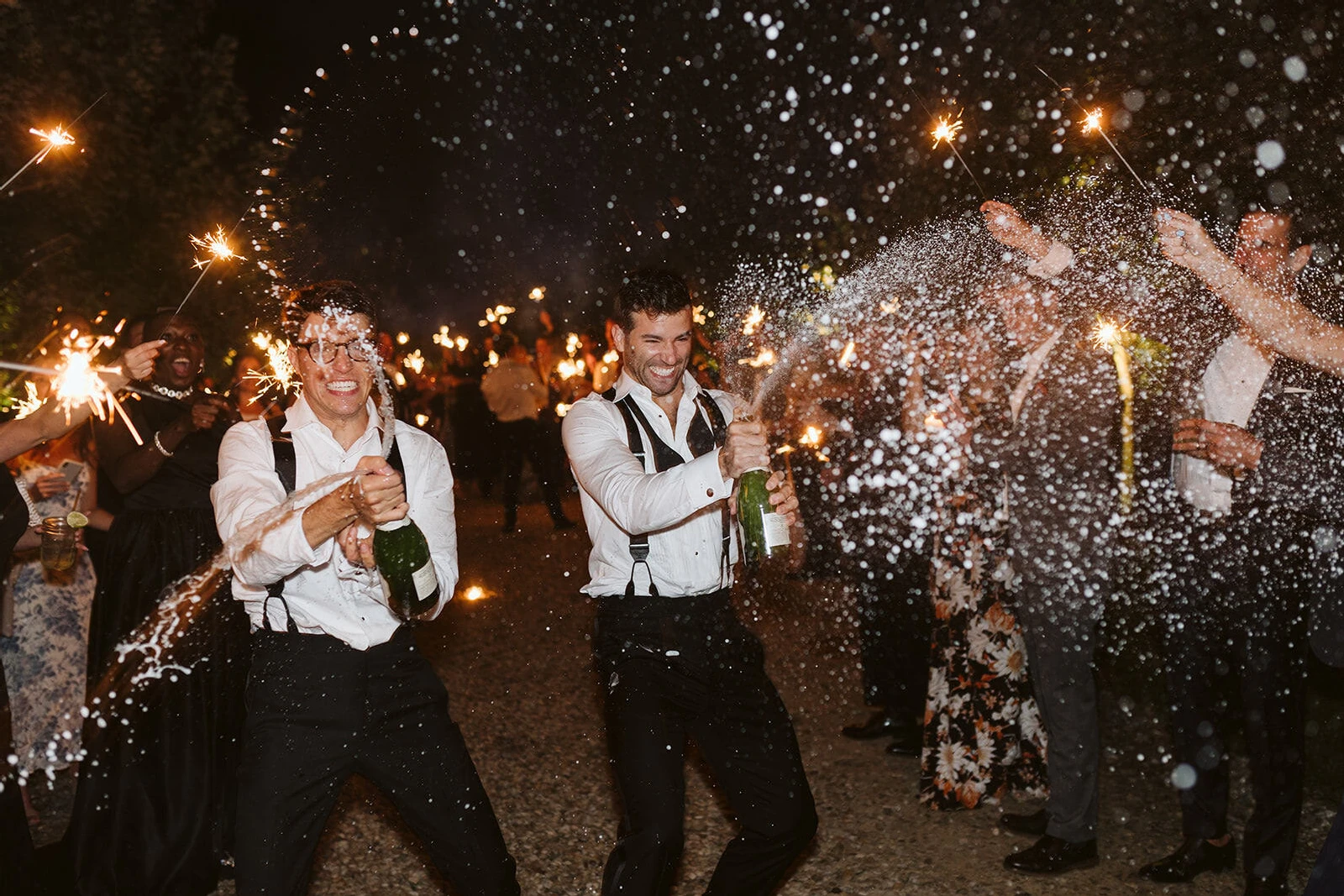 Holiday Wedding: Two grooms spraying champagne and smiling while loved ones surround them, cheering with sparklers.