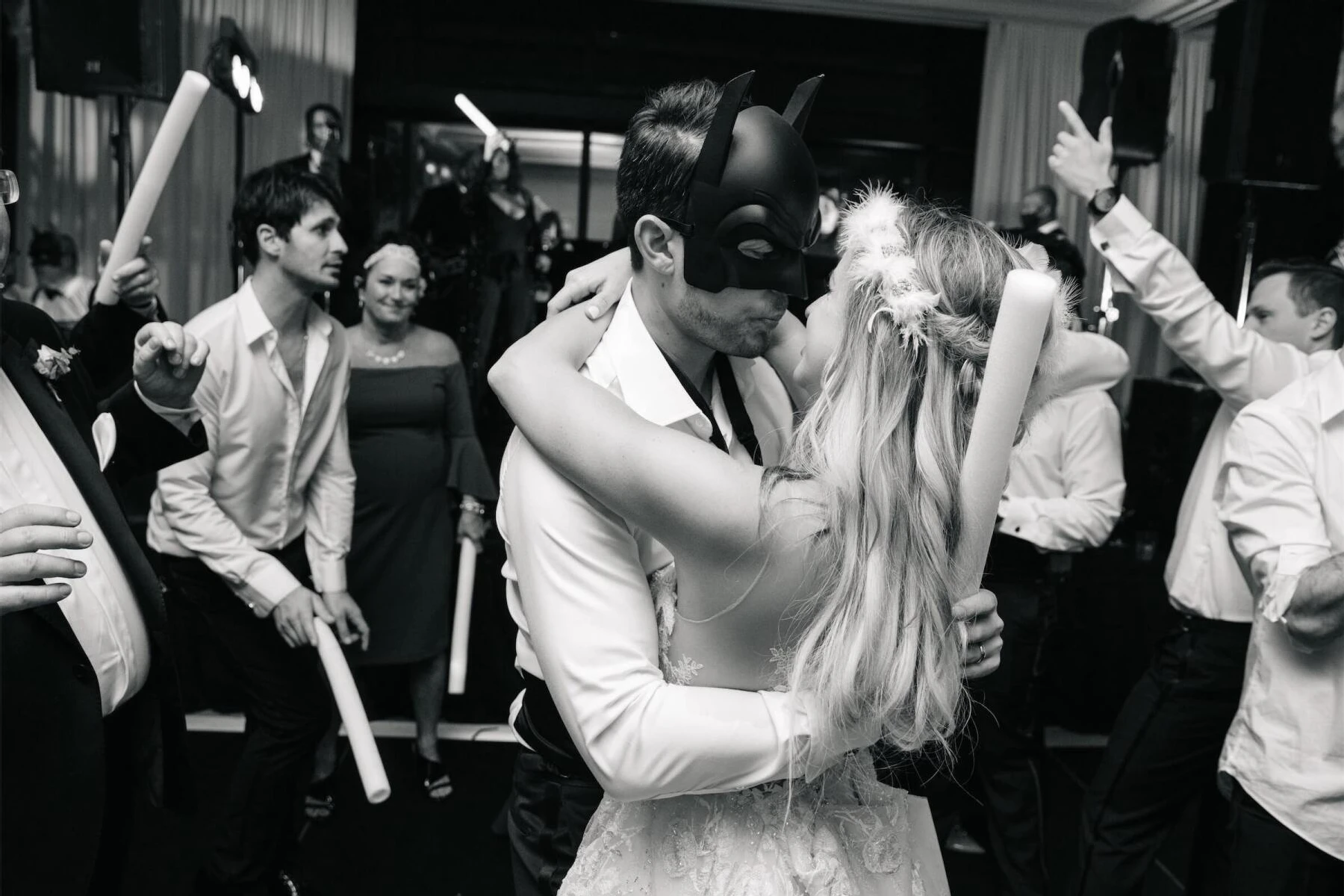 Holiday Wedding: A groom in a Batman mask and a bride wearing a flapper headpiece kiss on the dance floor while loved ones dancing around them.