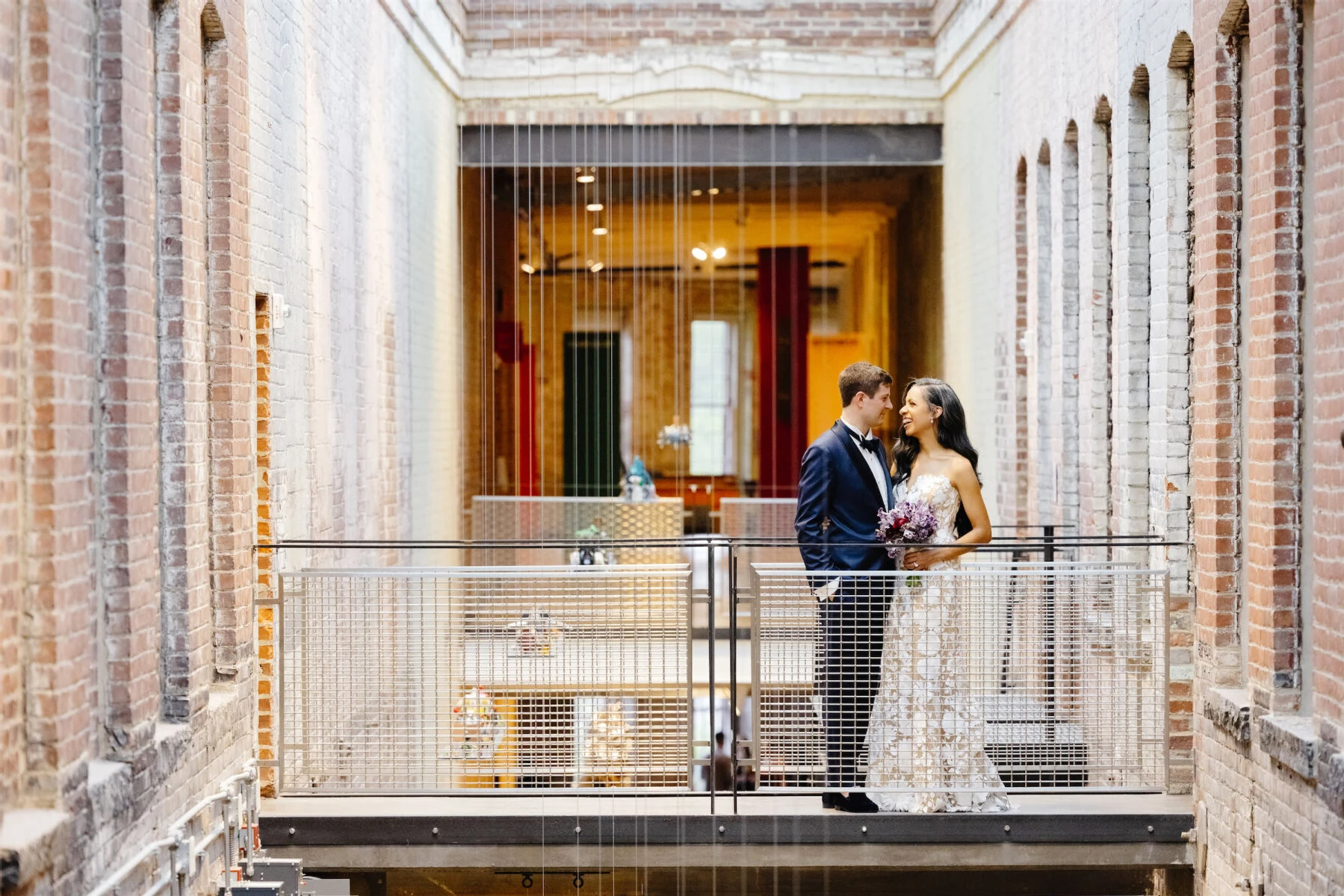 Industrial Wedding Venues: A bride and groom smiling at one another while standing on an industrial walkway with brick walls in the background at MASS MoCA.