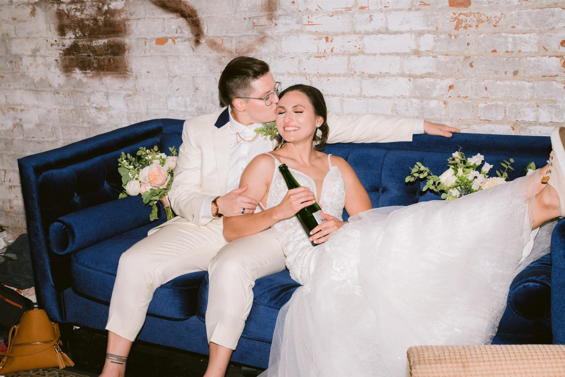 Industrial Wedding Venues: Two newlyweds snuggling on a blue couch, with one person kissing another on the cheek while the other holds a bottle of champagne and smiles.