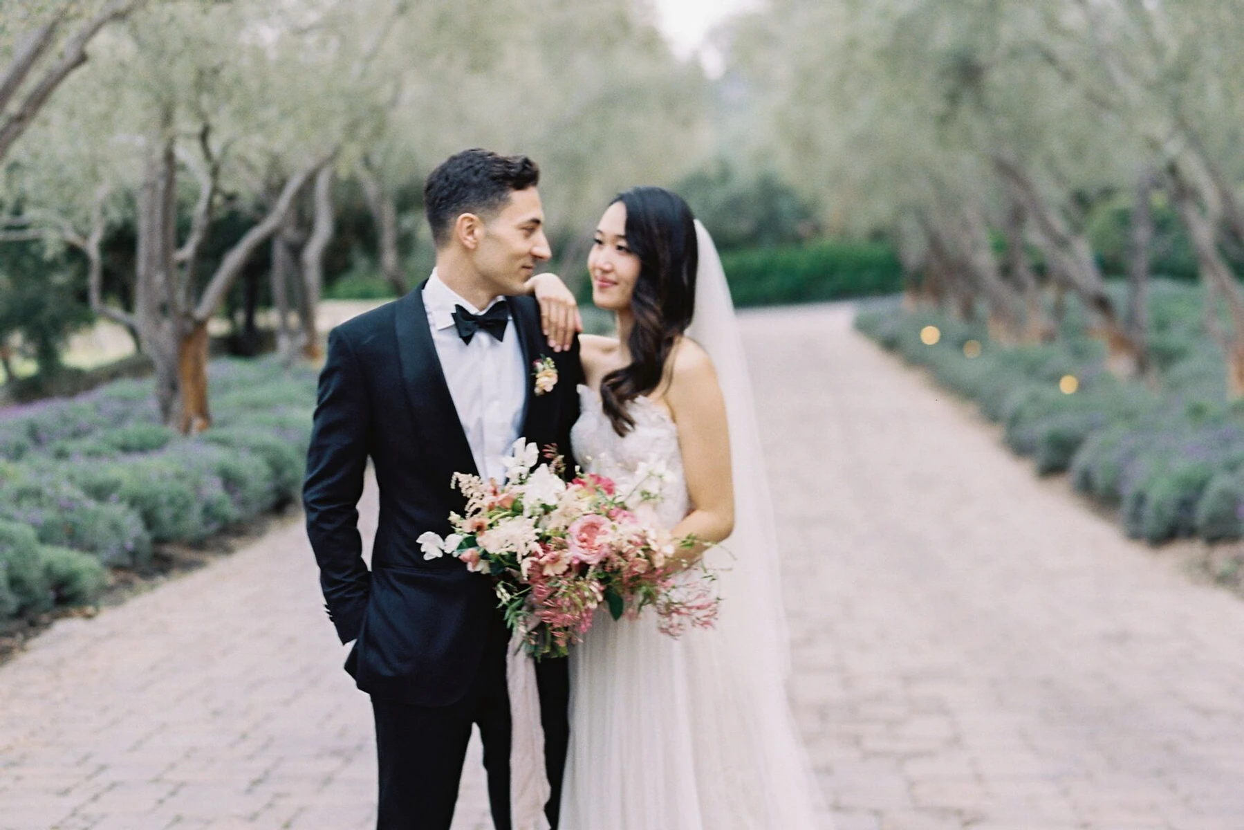 Groom in black tuxedo groom attire posing with bride on brick road