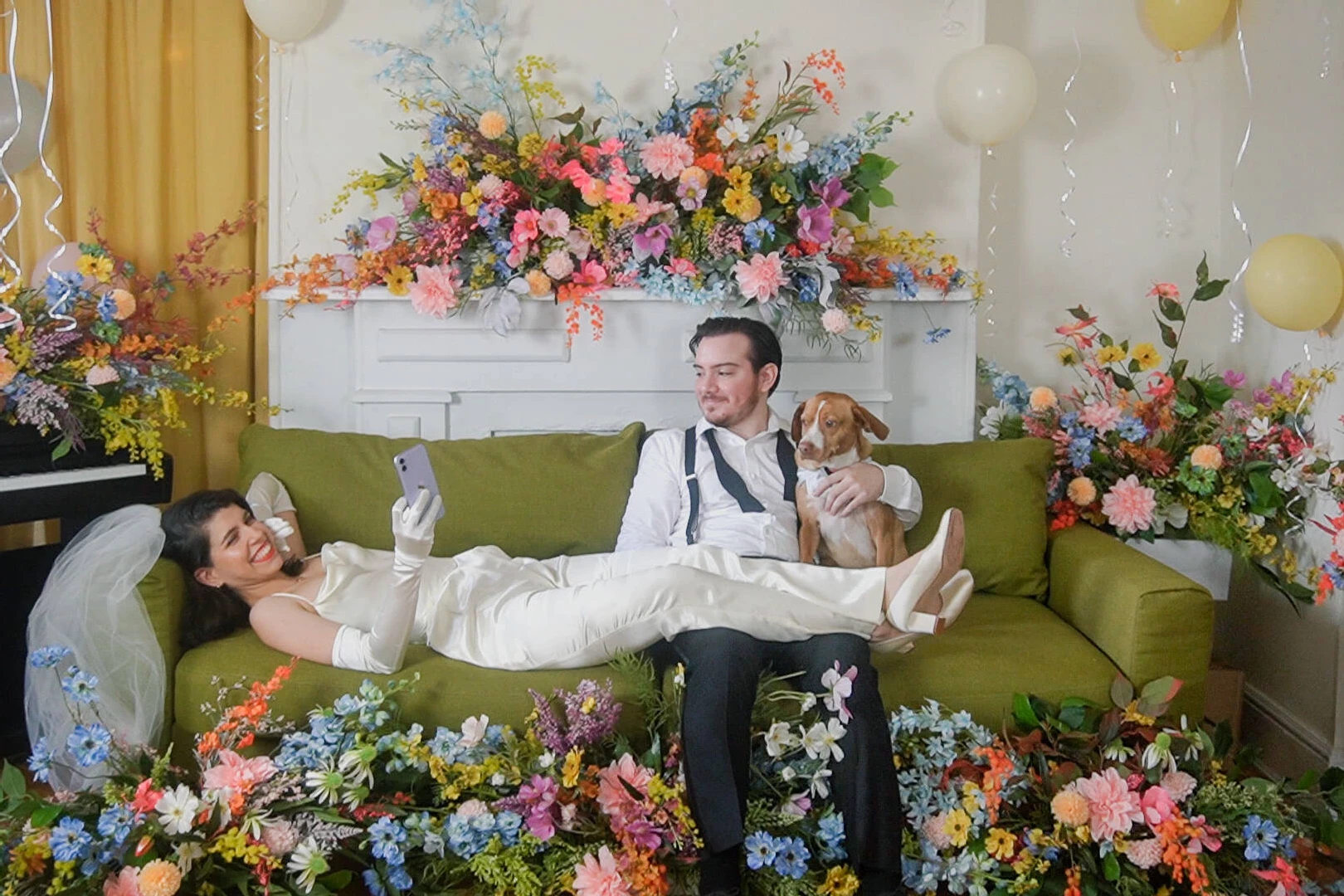 Latinx Heritage: A bride and groom sitting on a green couch with their dog, surrounded by flowers and balloons.