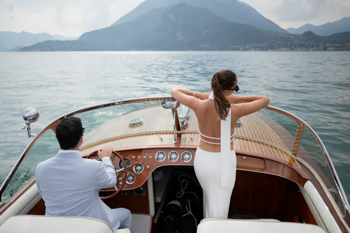 A backside view of a groom driving a boat on Lake Como in Italy, and the bride standing with her elbows resting on the boat's windshield.