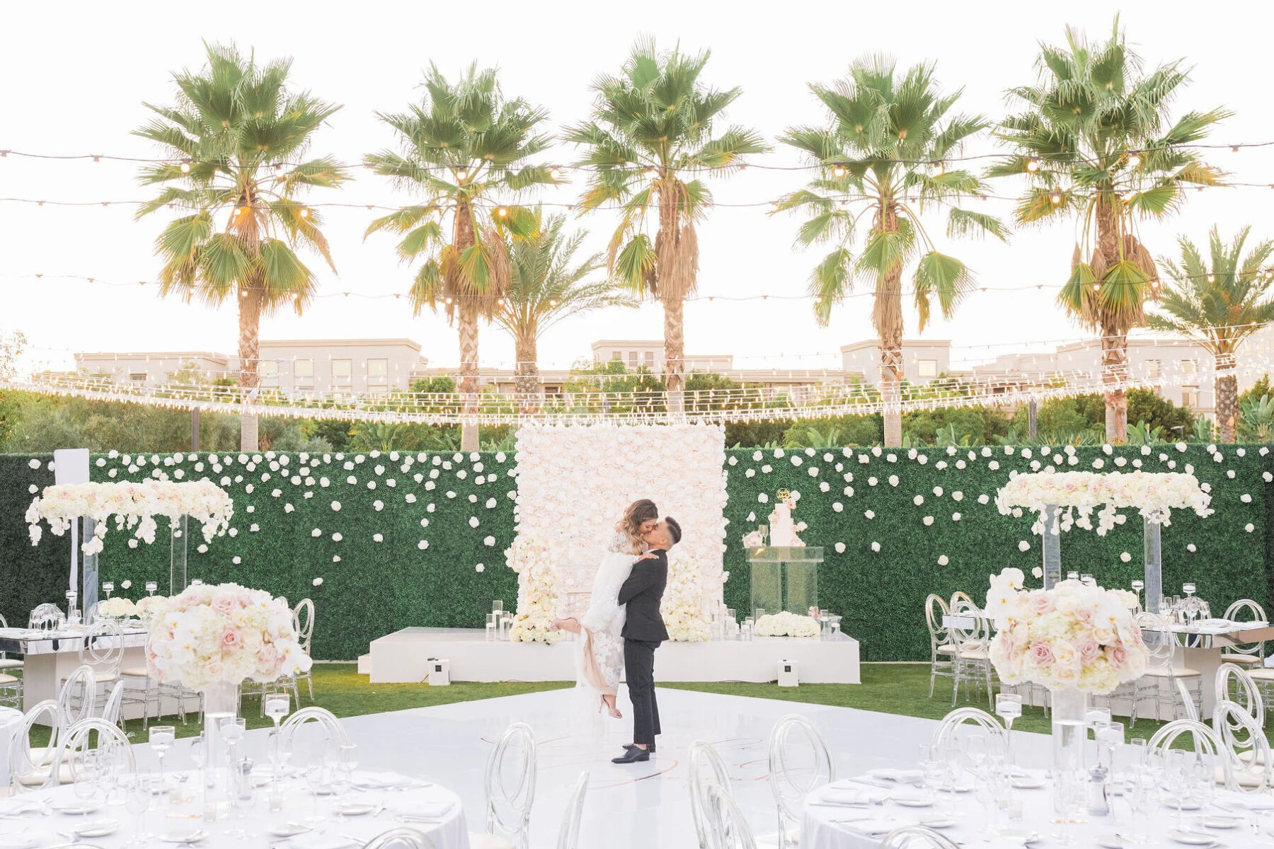 Marriott Weddings: A groom picking up a bride and kissing in an outdoor reception area covered in twinkle lights, white round tables and palm trees in the background.