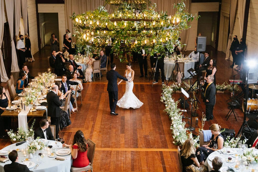 Pippin Hill Farm: A wedding couple dancing on a dance floor with an oversized chandelier covered in greenery and a band and guests watching around them.