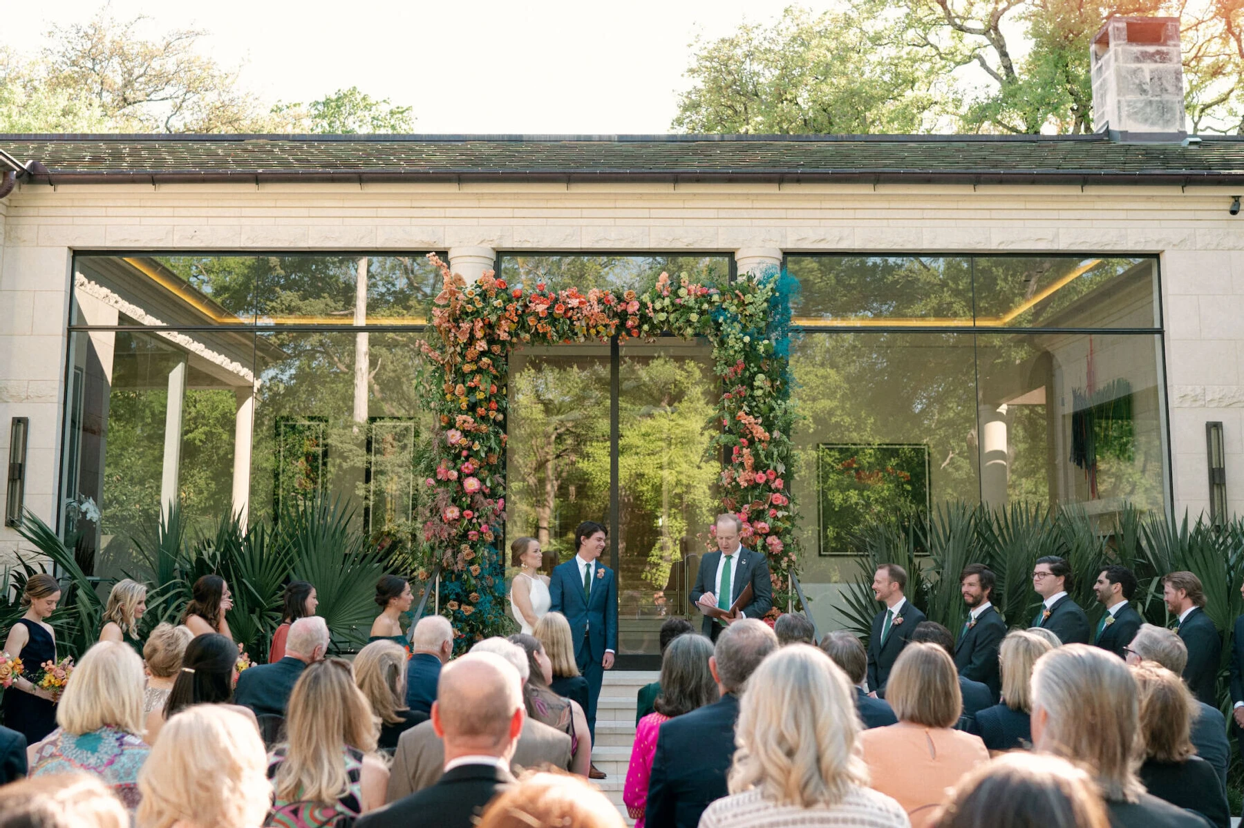 An outdoor wedding ceremony in front of a rainbow floral arch.