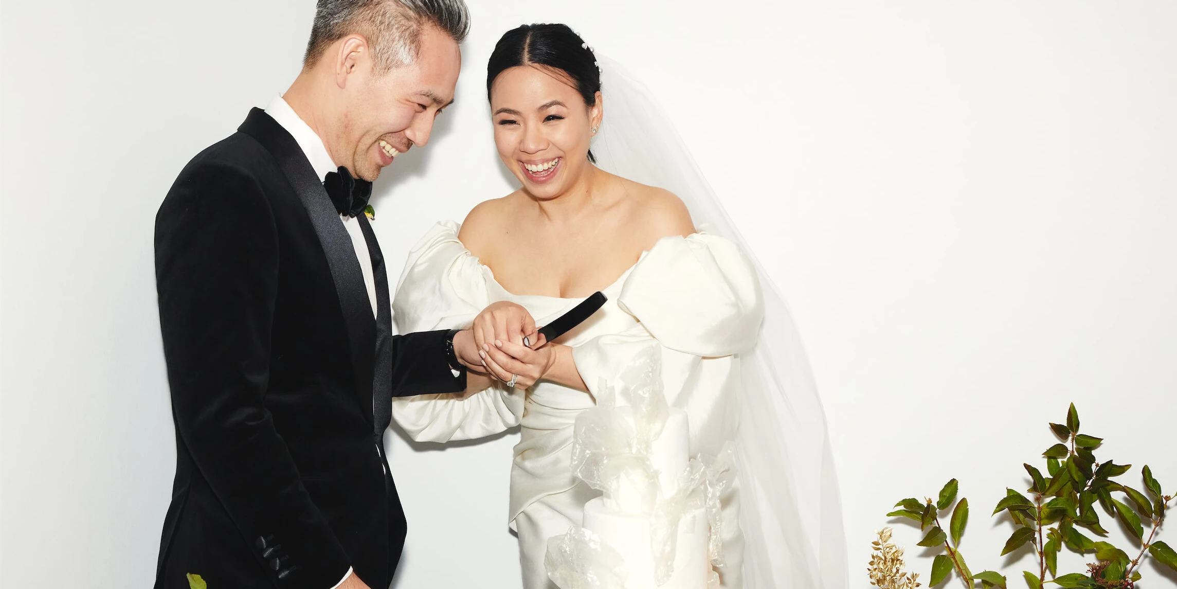 Wedding couple holding hands, laughing while cutting their modern white wedding cake together.