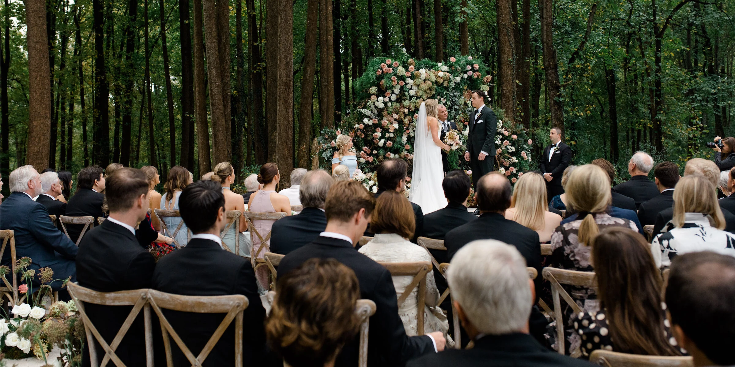 A rustic outdoor wedding ceremony in the woods included a floral backdrop behind the bride and groom.