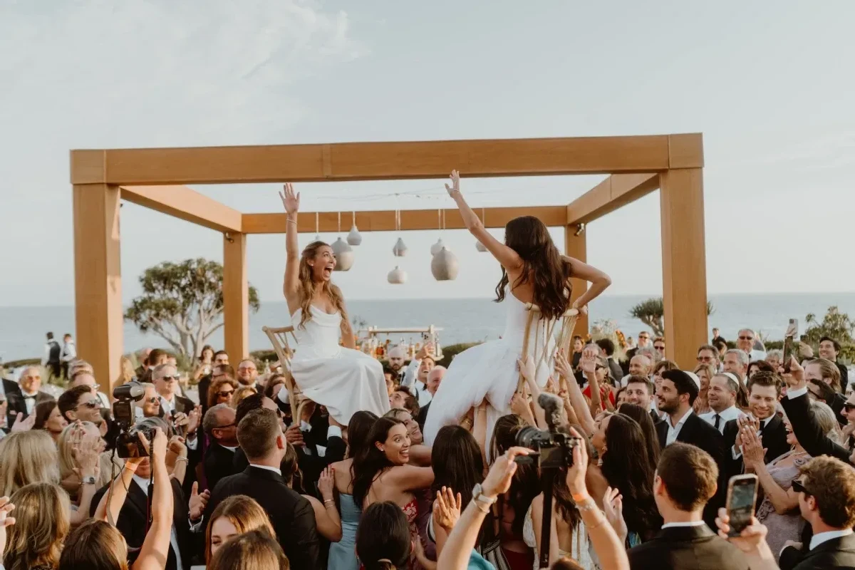 Two brides at an outdoor wedding reception in Laguna Beach, California, with guests surrounding them and holding them up on chairs.