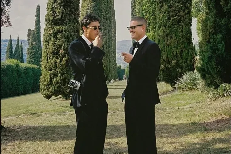 Two grooms in black suits with wide-leg trousers, standing among Italian cypress. 