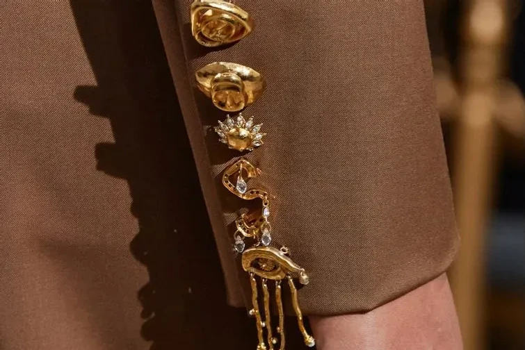 Brown suit sleeve with a row of gold surrealist rings and hardware set up the cuff, hand emerging at the bottom of the frame. 