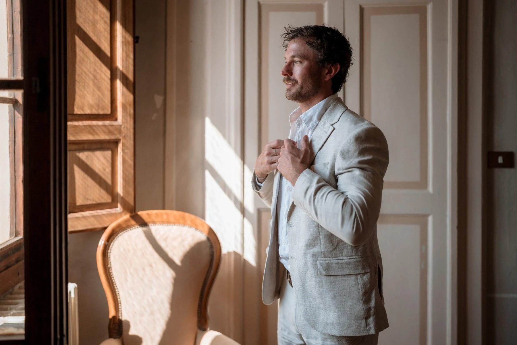 A groom looking out the window, wearing a beige linen suit. 