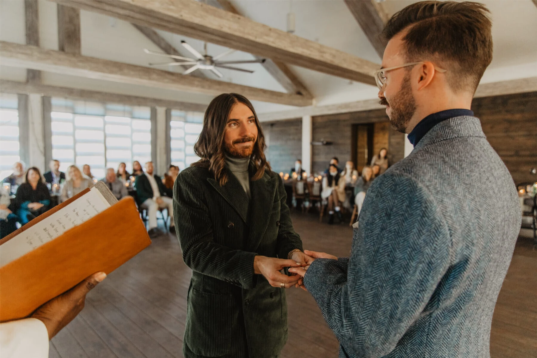 Wedding couple hold hands at the alter in front of their guests, who are seated at tables at their rustic barn venue.