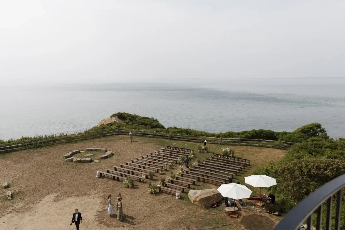 A drone image overlooking an outdoor Cape Cod ceremony setup with backless benches, with a view of the ocean in the distance.