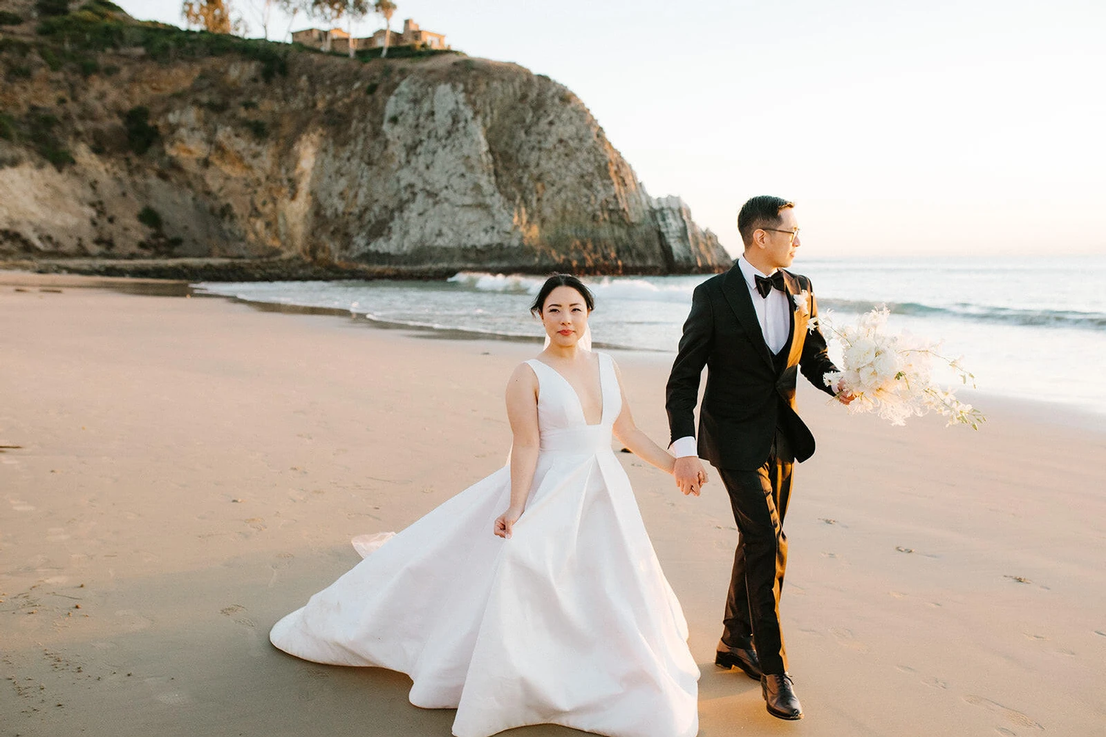 Wedding Dress Shopping: A bride and groom holding hands near a cliffside beach area in California.