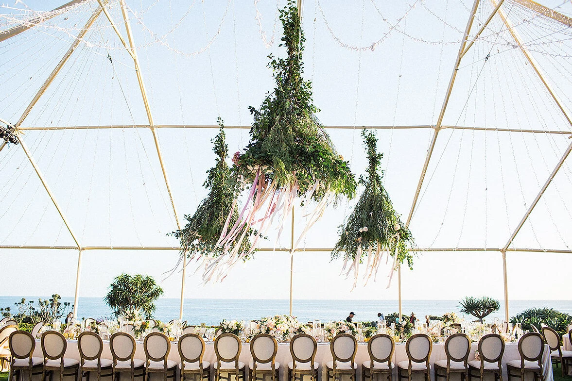 Wedding Etiquette Budget: Three oversize hanging floral installations over a long rectangular reception table at a tented wedding.