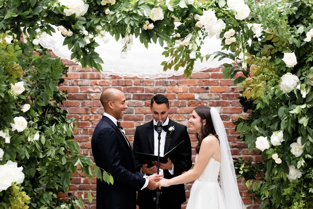 Wedding Website Examples: A wedding couple holding hands and smiling while their officiant speaks in front of a brick wall.