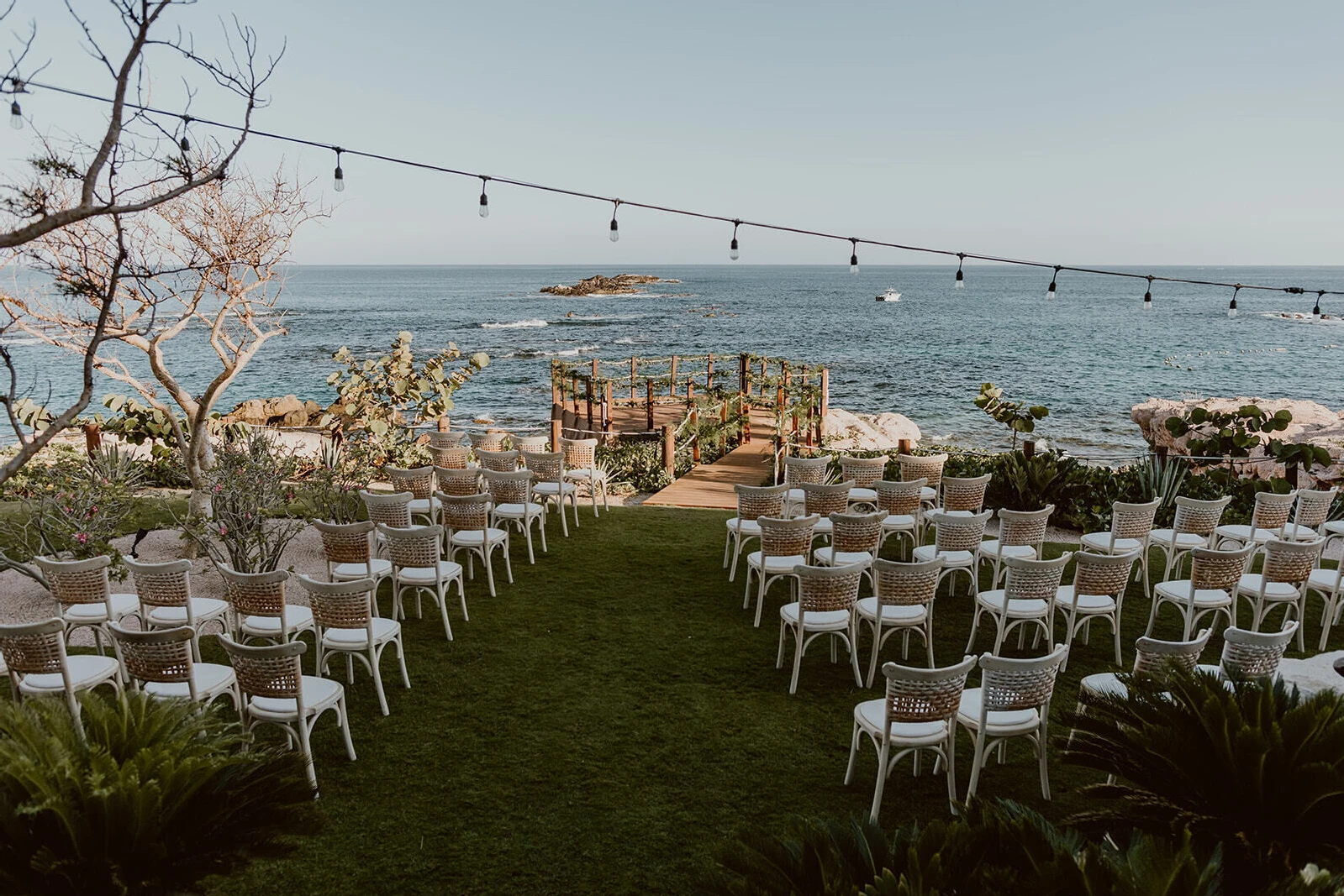 A waterfront ceremony setup with woven-back chairs and greenery as decoration for a Cabo wedding.