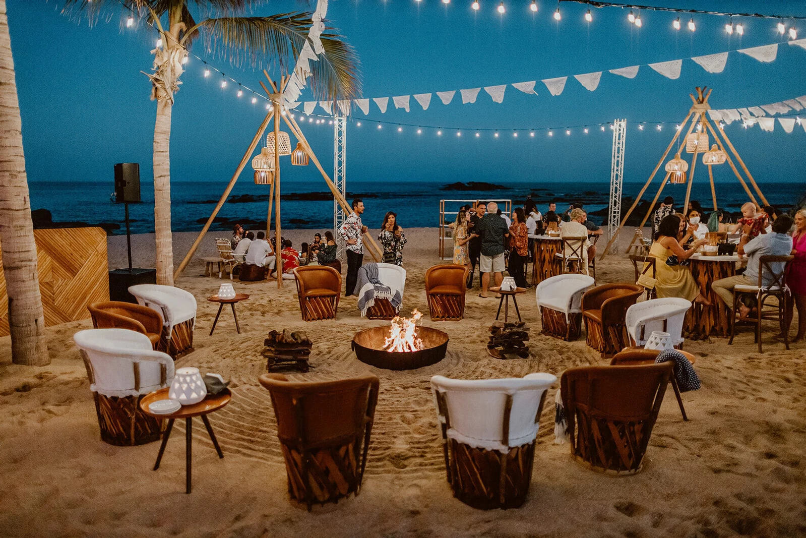 A welcome party on the beach in Cabo, with string lights and papel picado flags over a setup of various seating areas.