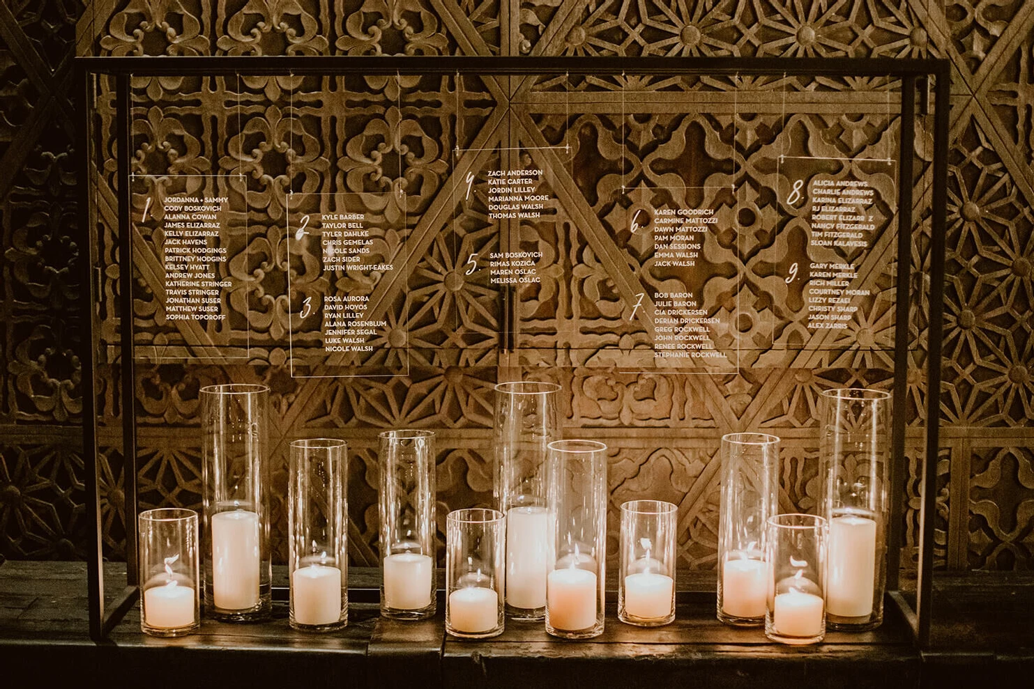 A hanging, clear seating chart is illuminated by pillar candles in glass.